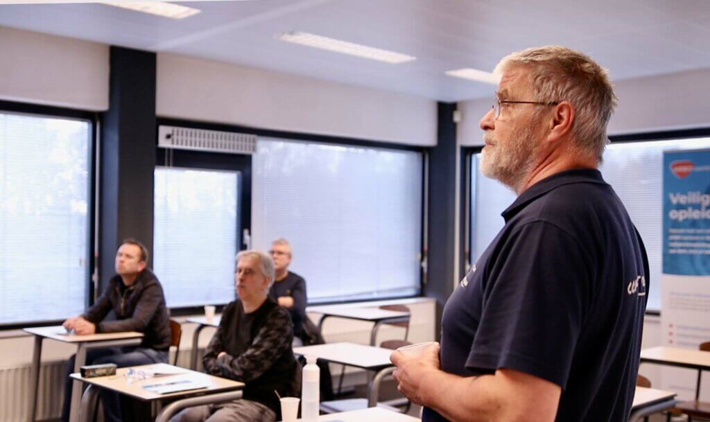 Een instructeur staat en deelt 3 tips bij het voorbereiden op een VCA cursus voor een klaslokaal met vier aandachtige mannen. De kamer, versierd met grote ramen, bureaus en een banner op de achtergrond, dient als de perfecte setting voor deze inzichtelijke sessie.