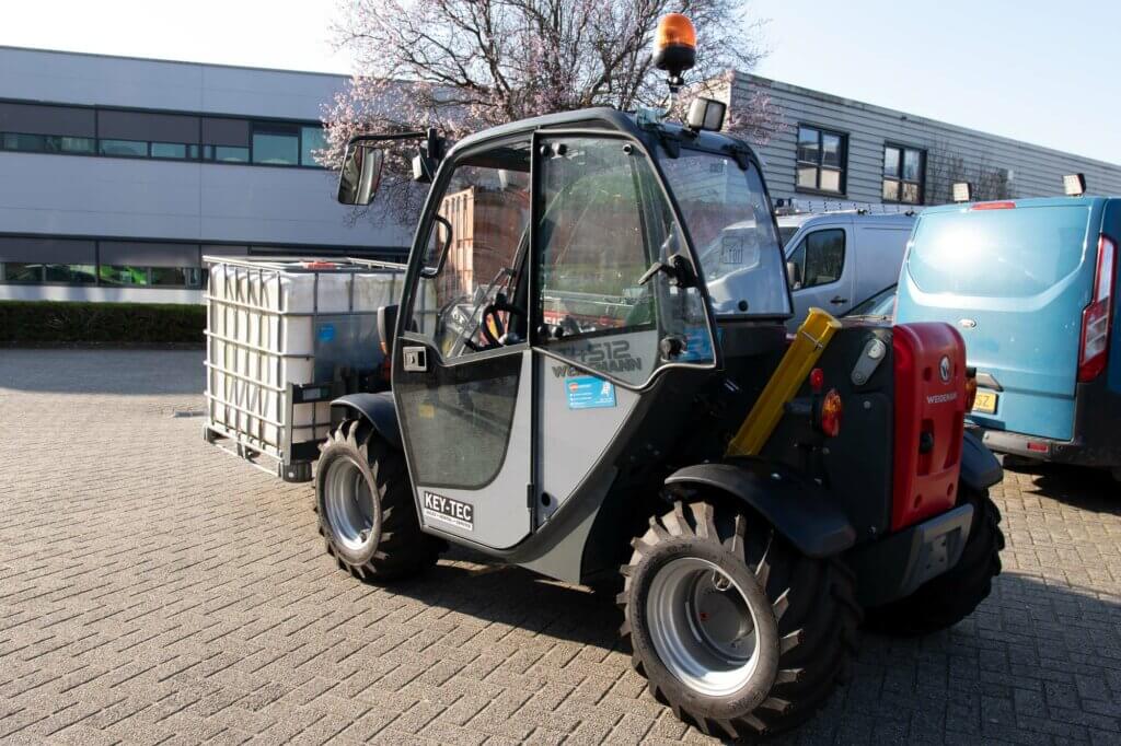 Een kleine vorkheftruck met een grote vloeistofcontainer staat geparkeerd op een verhard oppervlak in een industriegebied. Het heeft een afgesloten cabine en wordt verlicht door natuurlijk zonlicht. Op de achtergrond zijn moderne gebouwen en geparkeerde busjes te zien. In deze situaties gebruik je een verreiker voor verbeterde veelzijdigheid.
.