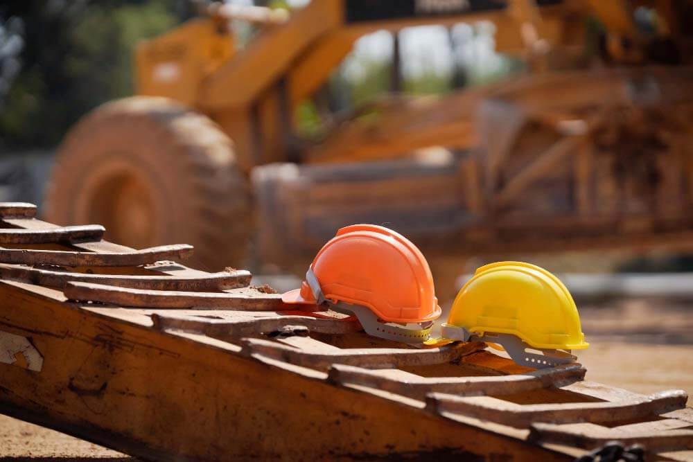 Twee bouwhelmen, een oranje en een gele, rusten op een stuk bouwmaterieel met grote wielen op de achtergrond. De scène weerspiegelt de uitdagingen waarmee een asfalteur wordt geconfronteerd als hij de risicos van een actieve bouwplaats onder fel daglicht navigeert.
