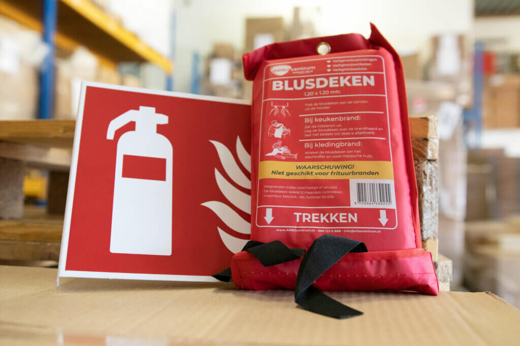 Een branddeken in rode verpakking staat op een tafel naast een brandblusserbord, met de nadruk op de 5 beste brandpreventietips. De achtergrond toont een magazijn met planken en dozen. De tekst op de verpakking en het bord is in het Nederlands.