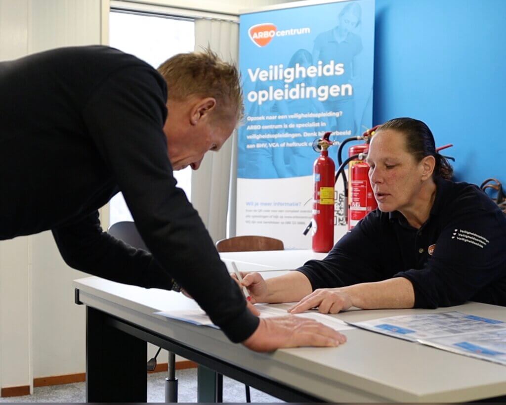 Een persoon in een zwart shirt leunt over een tafel en ondertekent een document, terwijl een andere persoon met lang haar naar het document wijst. Ze bevinden zich in een kamer met op de achtergrond een banner met Veiligheidsopleidingen, die illustreert waarom het volgen van een VCA-cursus bijdraagt aan een veiligere werkomgeving.