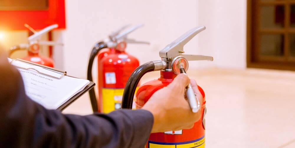 Een persoon die een rode brandblusser vasthoudt in een lichte kamer, omringd door anderen die in de rij staan voor inspectie. Met een klembord in de hand, kunnen ze tips voor het laten keuren van je brandblussers doornemen om veiligheid en naleving te garanderen.