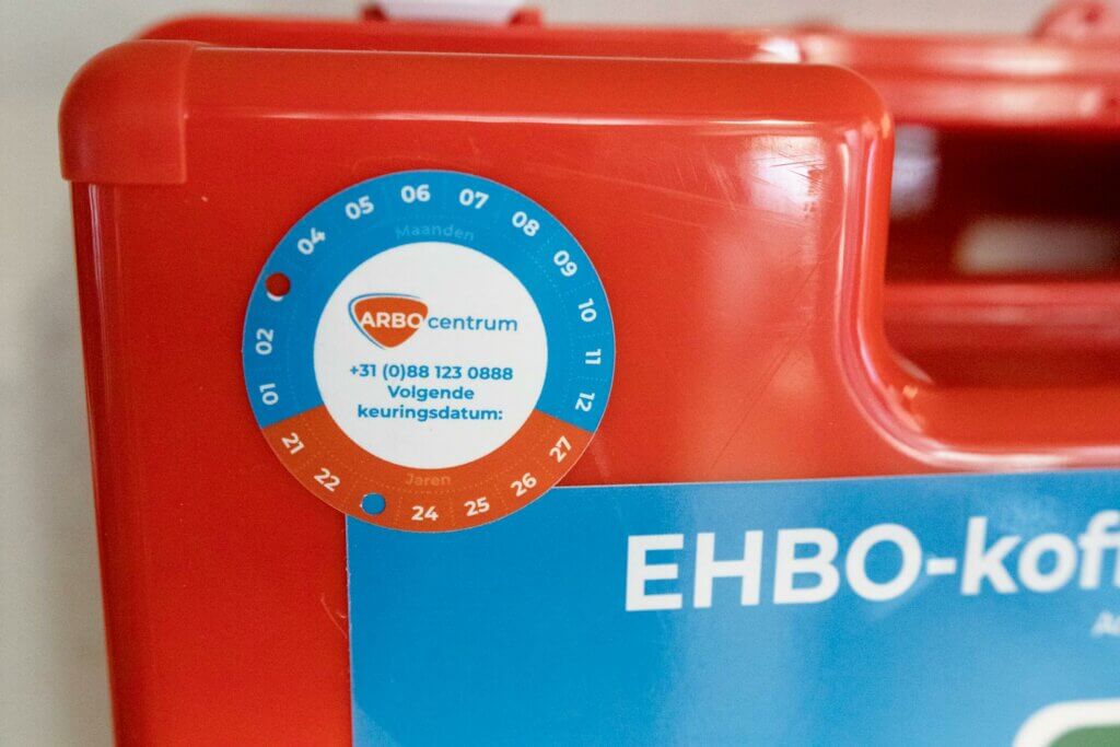 Close-up van een rode EHBO-doos met de aanduiding EHBO-koffer met een ronde keuringssticker van ARBOcentrum. De blauw-witte sticker geeft maanden en jaren weer en benadrukt het belang van regelmatige controles – de redenen waarom je zorgt voor onderhoud van EHBO koffers. Contactnummer is zichtbaar.