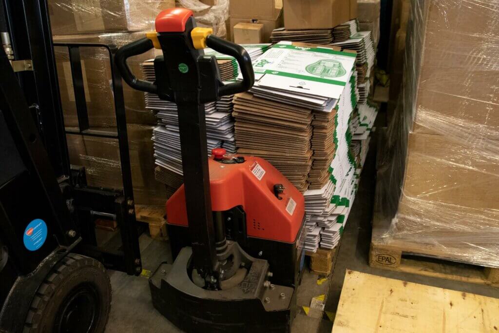 Een rode palletwagen staat geparkeerd in een magazijn naast een grote stapel bruine kartonnen platen—dit is waar een EPT voor dient. Op de achtergrond staan meerdere stapels dozen en een ingepakte pallet netjes gerangschikt, wat de efficiëntie van het materiaaltransport benadrukt.
