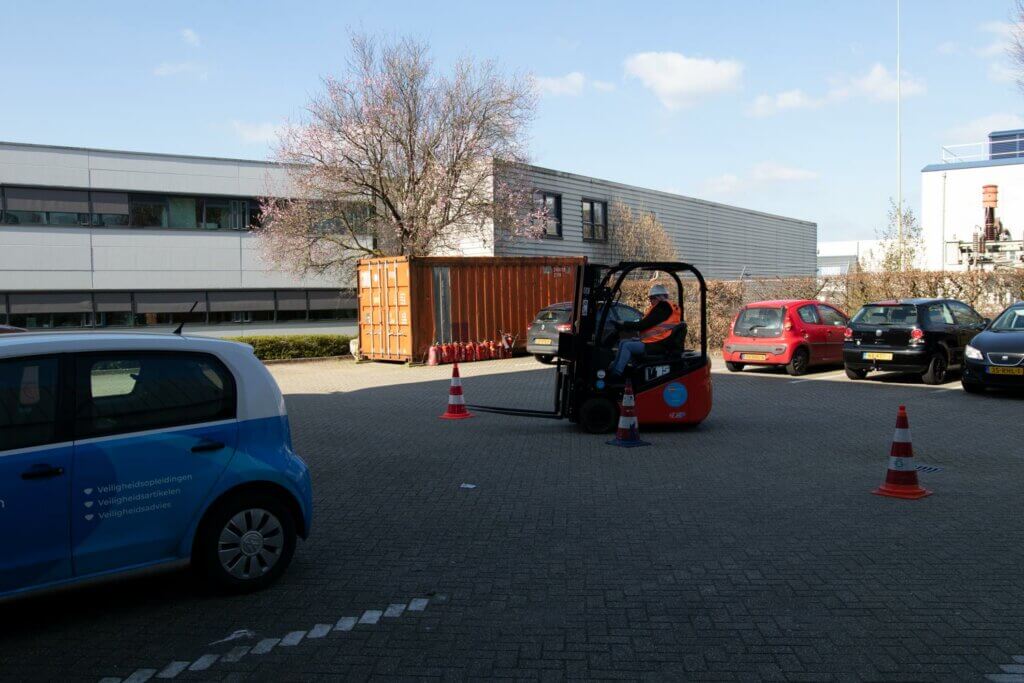 Een persoon bedient een vorkheftruck op een parkeerplaats omringd door veiligheidskegels, wat laat zien waarom een vorkheftruckcursus niet alleen belangrijk is, maar ook leuk. Met geparkeerde autos, een grote container en nabijgelegen gebouwen onder de zonnige hemel is het een boeiend tafereel.