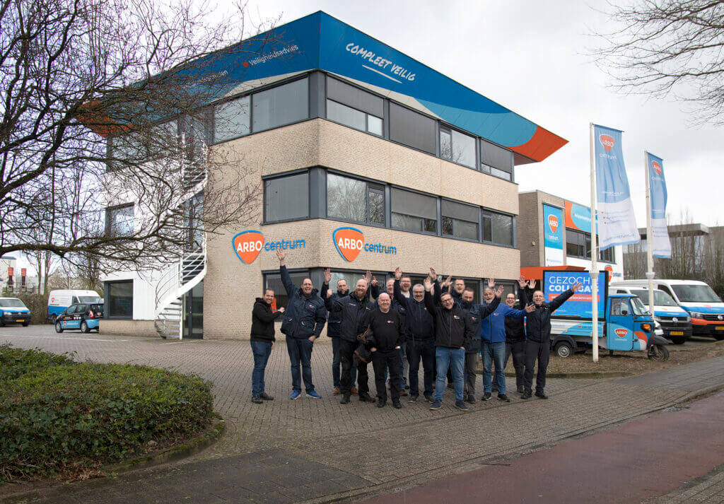 Een groep mensen staat buiten een modern gebouw met meerdere verdiepingen met de branding ARBO centrum. Ze lachen en steken hun armen omhoog. Op de achtergrond zijn verschillende bedrijfsbusjes en vlaggen met logos zichtbaar. De lucht is bewolkt.