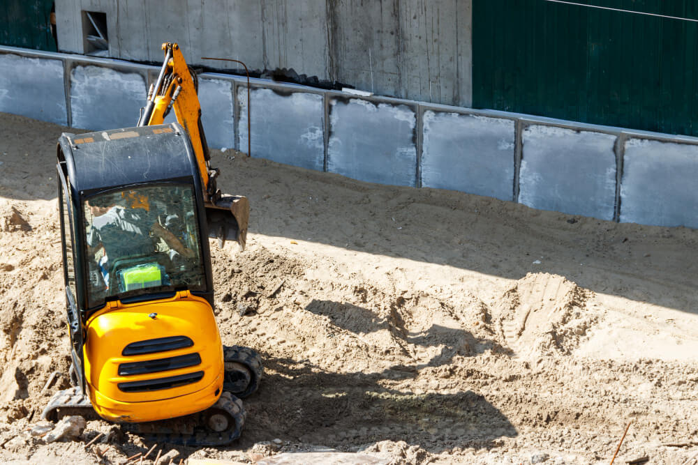 In deze situaties gebruik je een minigraafmachine: Een kleine gele graafmachine staat op een zanderige bouwplaats, vlak bij een betonnen muur. De arm van de graafmachine is uitgestrekt, klaar om te graven. De achtergrond bestaat uit gedeeltelijk geverfde groene oppervlakken en een grote betonnen constructie.