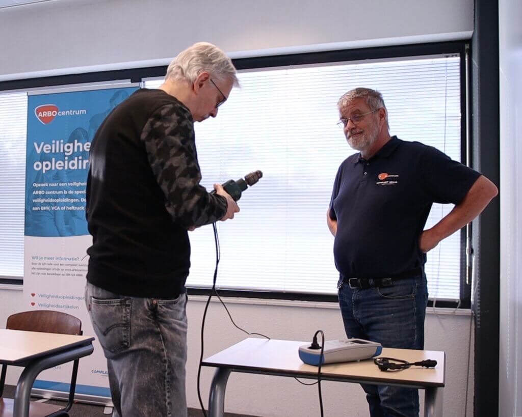 Twee mannen staan binnen bij een raam met elkaar te praten. De ene man houdt een boormachine vast, gefocust op zijn taak, terwijl de ander met zijn handen op zijn heupen aandachtig toekijkt. Achter hen hangt een spandoek van een opleidingscentrum waarop staat hoe een NEN 3140-cursus de kansen op de arbeidsmarkt vergroot. Op de voorgrond staan tafels.