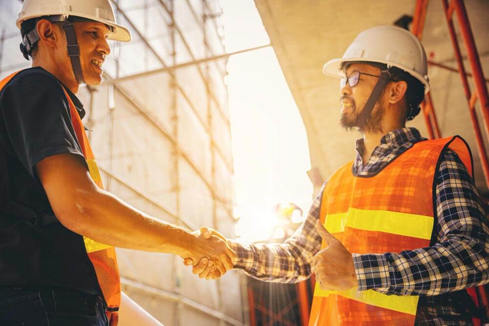 Twee bouwvakkers met helmen en oranje veiligheidsvesten schudden elkaar de hand op een bouwplaats. Een arbeider, die zijn goedkeuring geeft met een duim omhoog, benadrukt de redenen waarom elk bedrijf een RI&E zou moeten hebben. Steigers en zonlicht omlijsten het tafereel op de achtergrond.