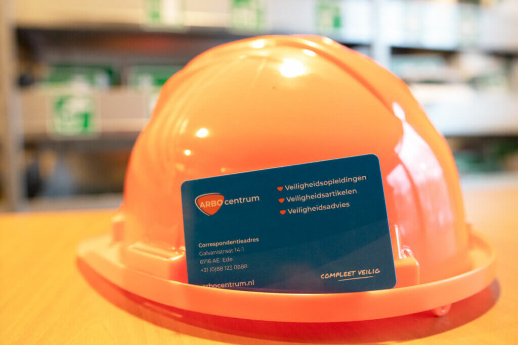 Een roze helm rust op een houten oppervlak en bedekt gedeeltelijk een blauw visitekaartje met tekst. Op de wazige achtergrond hangen planken met groene veiligheidsborden, die misschien wijzen op het belang van het behalen van uw VIL-VCU-diploma en drie dwingende redenen om dit te overwegen.