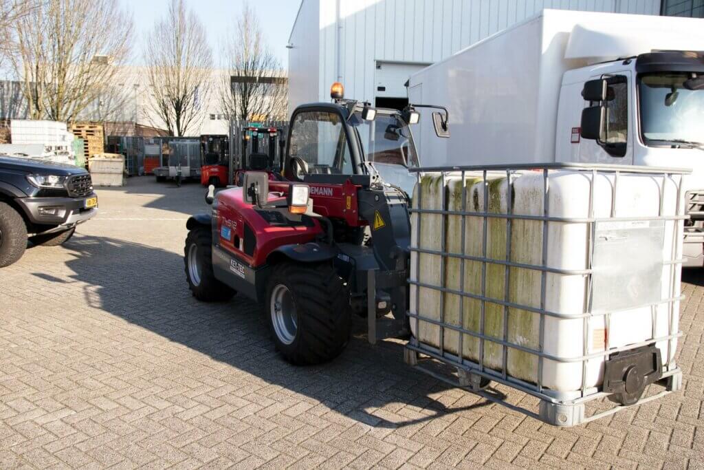 Een rode vorkheftruck, die de voordelen van een starre verreiker laat zien, draagt een grote container gevuld met gelige vloeistof en staat buiten geparkeerd op een zonnige dag. Industriële gebouwen en andere voertuigen vormen de perfecte achtergrond voor deze demonstratie van robuuste efficiëntie.