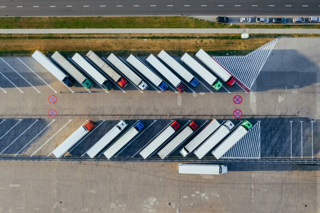 Luchtfoto van een parkeerplaats met meerdere vrachtwagens die diagonaal geparkeerd staan, elk op een aangewezen plek. De parkeerplaats heeft gemarkeerde parkeerverbodszones met grote rode Xen en grenst aan een weg met meerdere rijstroken bovenaan de afbeelding, perfect om 3 manieren om code 95-voorschriften te bekijken tijdens rustplaatsen.
