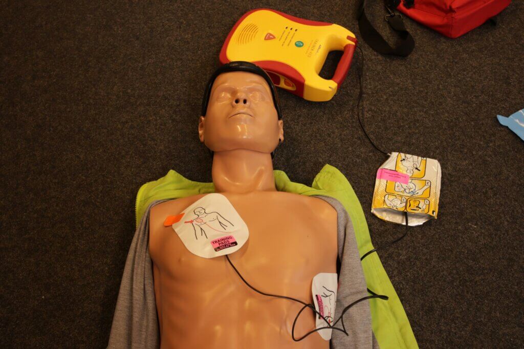 Een reanimatie-oefenpop ligt op de grond met zelfklevende elektrodepads op de borst. Een defibrillator is boven het hoofd geplaatst, gedeeltelijk bedekt met een groene doek op de schouders—dit is waarom iedereen een BHV-cursus zou moeten volgen.