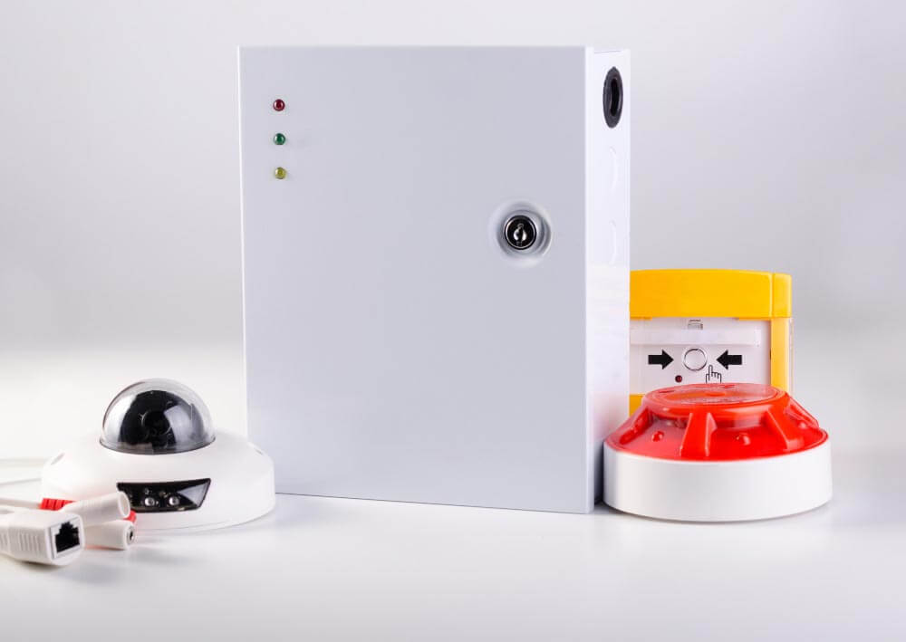 Een rechthoekig wit beveiligingsapparaat met indicatielampjes en een sleutelgat wordt geflankeerd door een witte domecamera aan de linkerkant en een geel en rood alarmsysteem, essentieel voor de taken van een beheerder brandmeldinstallatie, aan de rechterkant. Ethernetkabels zijn gedeeltelijk zichtbaar op de voorgrond.