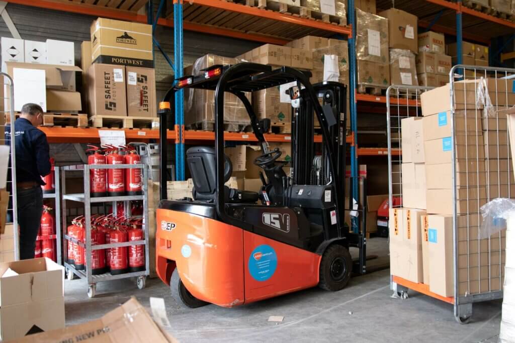 In een drukke magazijnscène hint een kleine oranje vorkheftruck naar de mogelijkheden met een vorkheftruckcertificaat. Hij staat geparkeerd bij planken vol dozen, terwijl een arbeider in een blauw jasje efficiënt beweegt. Rode brandblussers staan bij elkaar op de voorgrond, terwijl er rechts nog meer dozen netjes zijn opgestapeld.