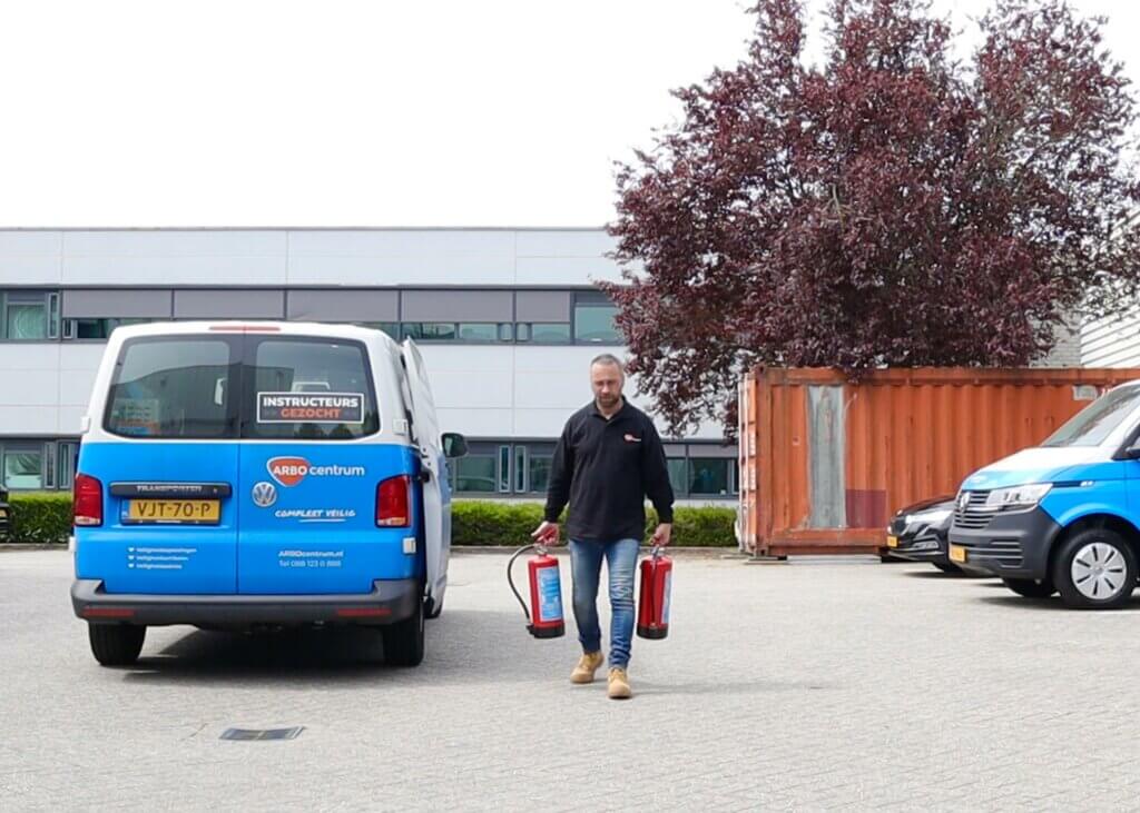 Een persoon die over een parkeerplaats loopt, met twee brandblussers in zijn handen, is een voorbeeld van waarom iedereen een BHV-cursus zou moeten volgen. Een blauwe bestelwagen met het opschrift INSTRUCTEURS staat vlakbij geparkeerd, naast een ander voertuig en een zeecontainer, met een boom en gebouw als achtergrond.
