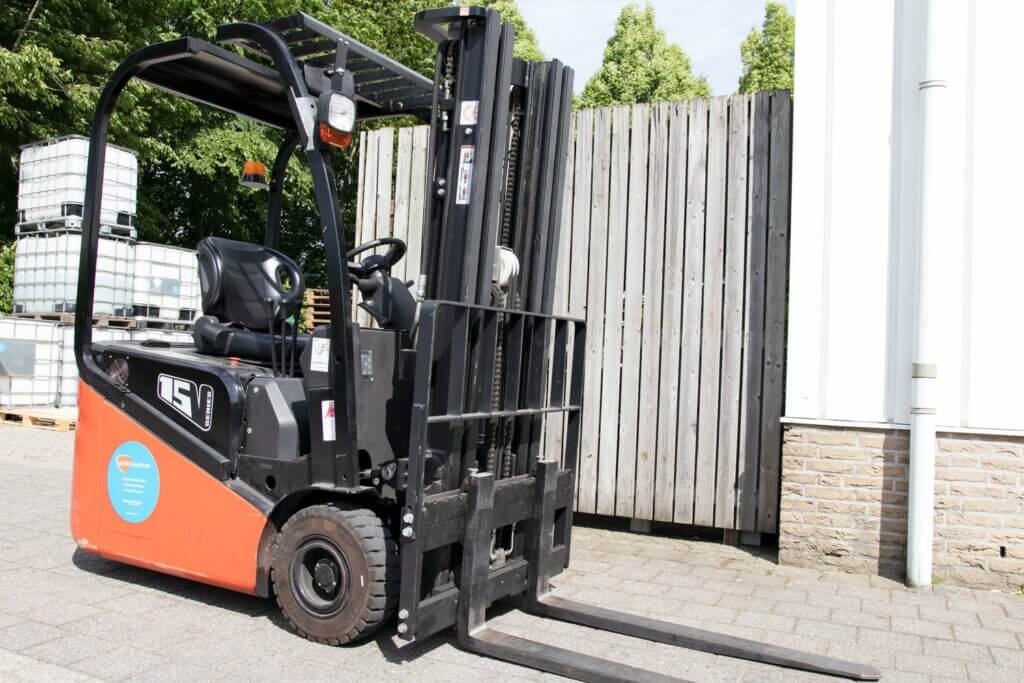 Een zwart-oranje heftruck staat geparkeerd op een geplaveid terrein bij een houten hek en een wit gebouw. De vorken zijn naar voren gericht, klaar voor actie. Op de achtergrond zijn opgestapelde plastic containers zichtbaar, die benadrukken waarom de opleiding tot heftruckchauffeur volg je bij ons om 3 redenen.