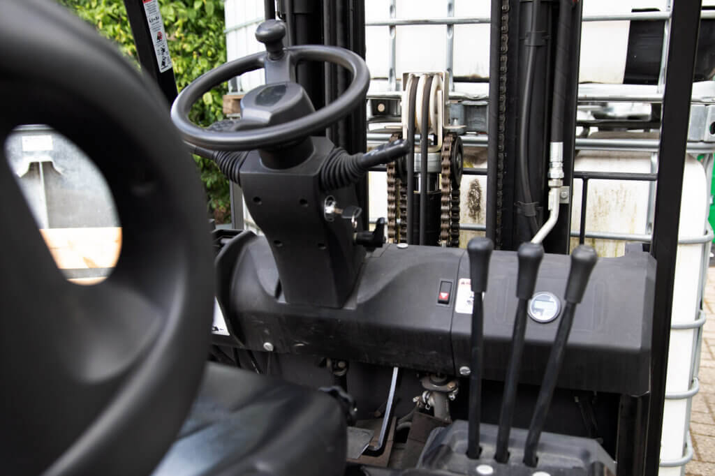 Close-up van het bedieningsgedeelte van een vorkheftruck, met de nadruk op het stuur, de hendels en de versnellingen. Op de industriƫle achtergrond zijn zichtbare containers te zien. Inschrijven voor een heftruckcursus? Dit zijn de opties die je nodig hebt om je vaardigheden in deze dynamische omgeving te verbeteren.