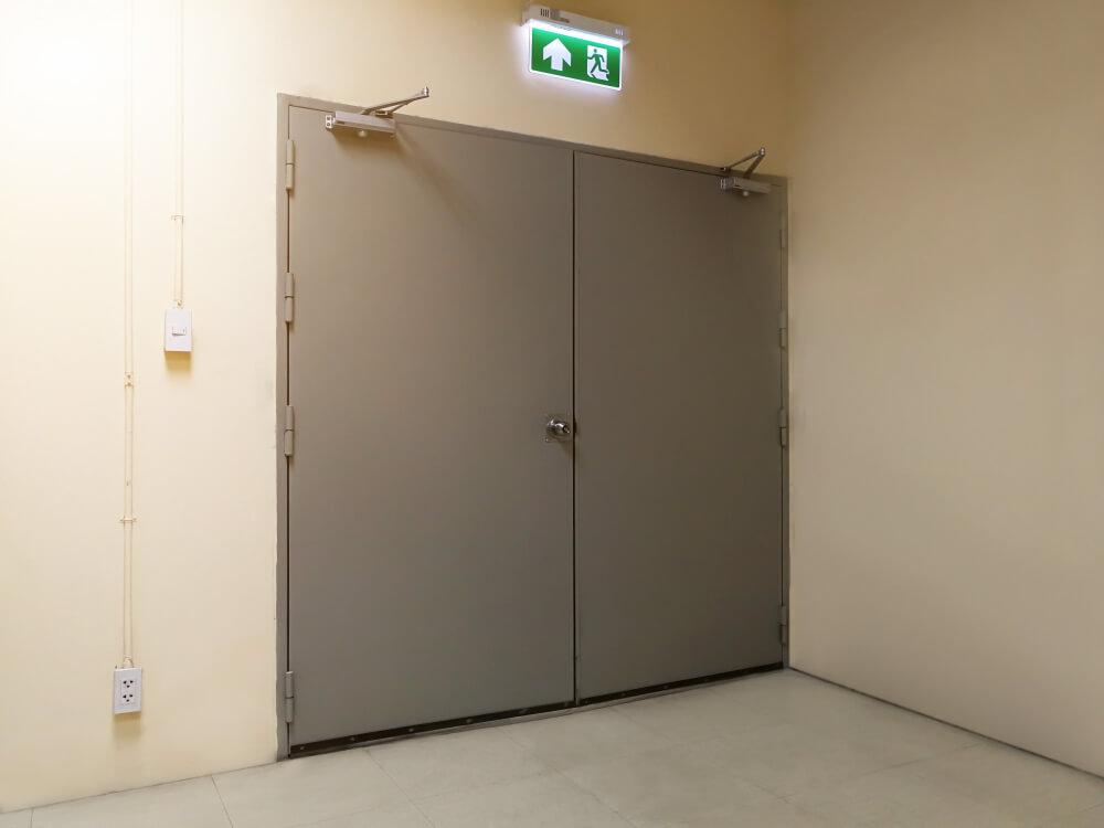 Een gesloten grijze metalen deur in een beige gang hint naar veiligheid met een groen exit-bord erboven, met een rennende figuur en een pijl omhoog. De eenvoudige muren en vloer zorgen voor eenvoud, terwijl er een stopcontact op de linkermuur rust. Je hebt een ontruimingsplan: dit is hoe het werkt voor snelle evacuatie.