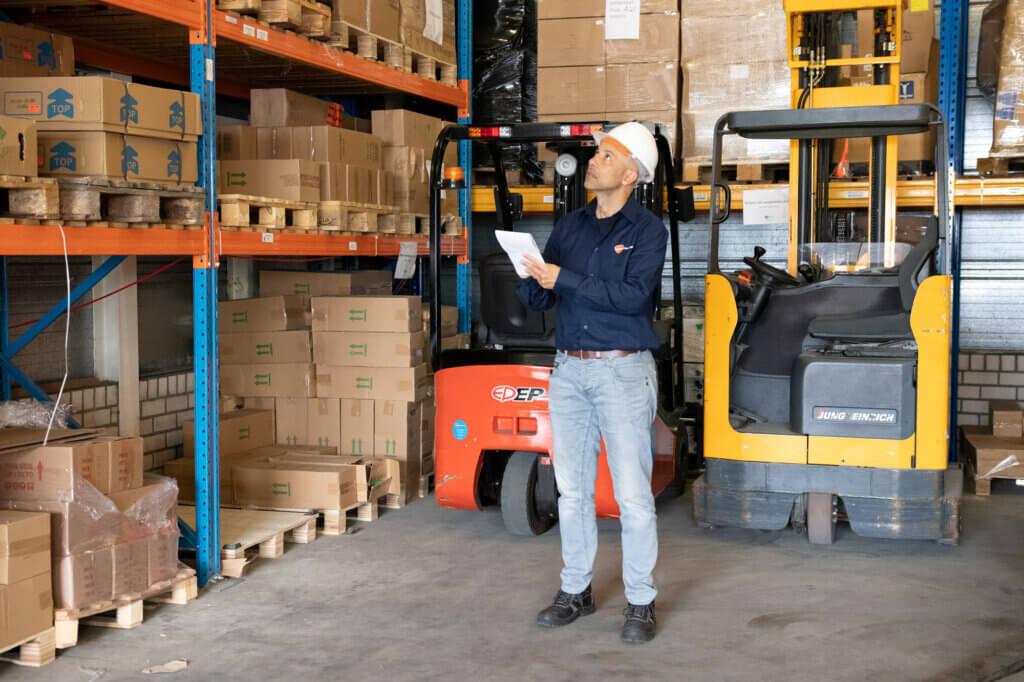 Een man met een witte helm en een blauw shirt houdt een stuk papier vast met het label Risico Inventarisatie en Evaluatie terwijl hij in een magazijn staat. Om hem heen staan dozen op planken, wat onderstreept waarom zulke evaluaties cruciaal en verplicht zijn. In de buurt staan twee vorkheftrucks geparkeerd.