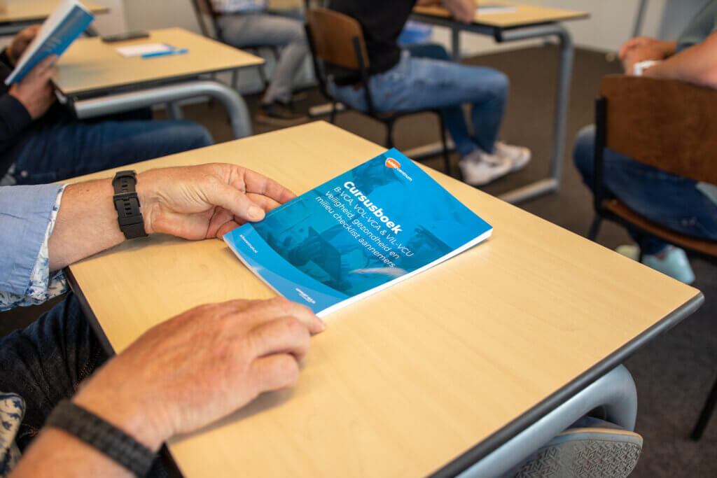 Close-up van een persoon die een blauwe gids met de titel Gas Book vasthoudt op een bureau in een klaslokaal. Andere studenten in casual kleding zitten aan lichte houten bureaus, sommigen houden soortgelijke boekjes vast. Deze scène is een voorbeeld van hoe de VCA-controle werkt in onderwijsomgevingen.