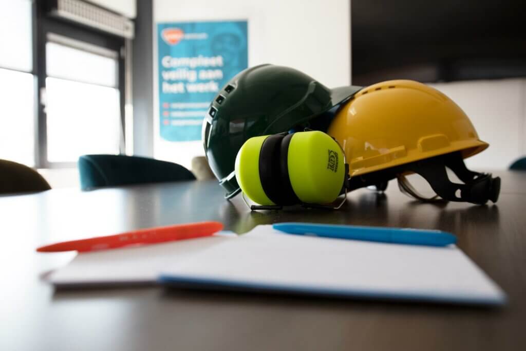 Een gele en groene bouwhelm, samen met groene oorkappen, staan op een houten tafel. Op de voorgrond suggereren een rode pen, blauwe pen en een leeg notitieboek het maken van aantekeningen voor het selecteren van welke VCA cursus past het best bij jou. Een veiligheidsposter is wazig op de achtergrond.