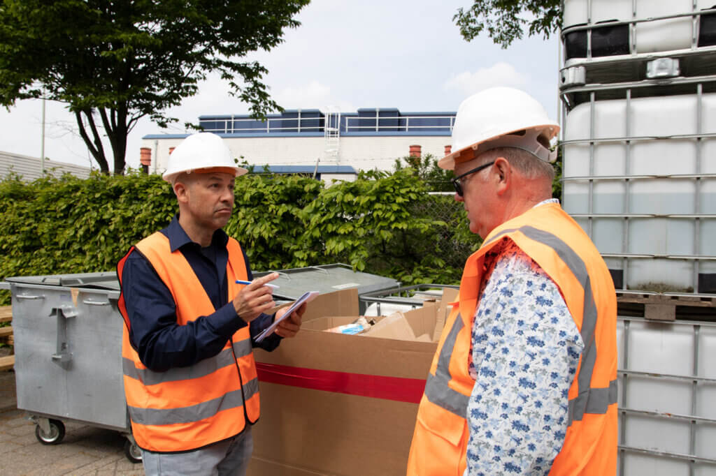 Twee mannen in veiligheidsvesten en helmen staan buiten bij industriële containers. Een van hen houdt een notitieblok vast, schijnbaar aantekeningen makend, wat de groeiende vraag naar Het VCA-diploma weerspiegelt. Groen en industriële apparatuur vormen de achtergrond.