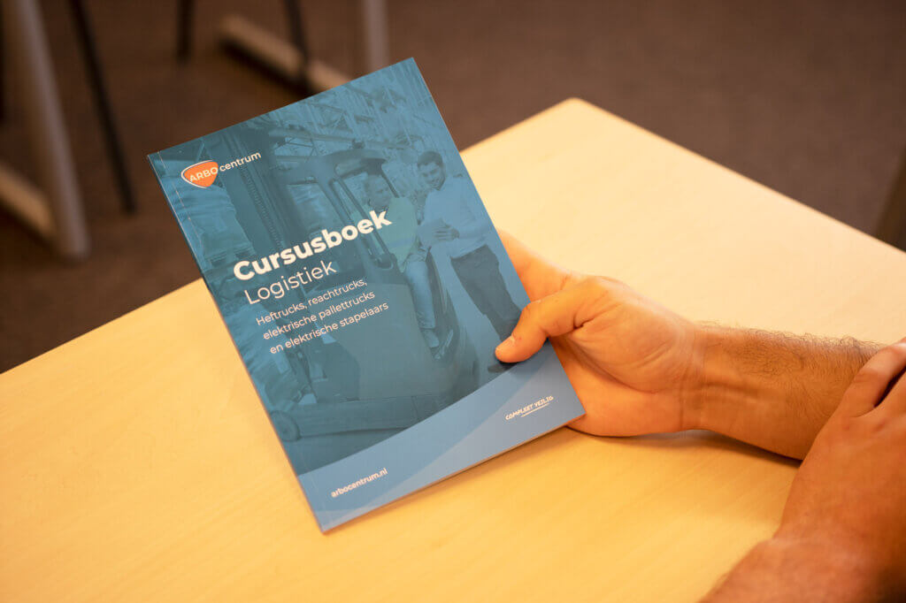 Een persoon houdt een blauw boek vast met de titel Cursusboek Logistiek in het Nederlands, gericht op netwerklogistiek, kostenbeheersing, productie en duurzame aanpassingen - een essentiële bron als u van plan bent om uw heftruckcertificaat te halen. Het boek ligt op een houten tafel.