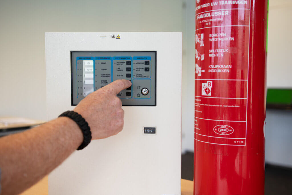 Een persoon wijst naar een bedieningspaneel op een witte doos, mogelijk een brandalarmsysteem, naast een grote rode brandblusser. Het paneel toont knoppen en indicatoren, vergelijkbaar met die welke je zou bestuderen in de NEN 3140-cursus. Een armband is zichtbaar om de pols van de persoon.