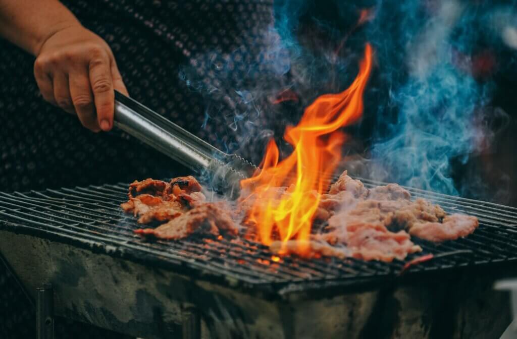 Een persoon grilt vlees op een barbecue, waarbij hij met een tang stukken omdraait op een metalen rooster. Feloranje vlammen stijgen op tussen het vlees en blauwe rook dwarrelt eromheen. Het grillen gebeurt buiten in een informele setting.