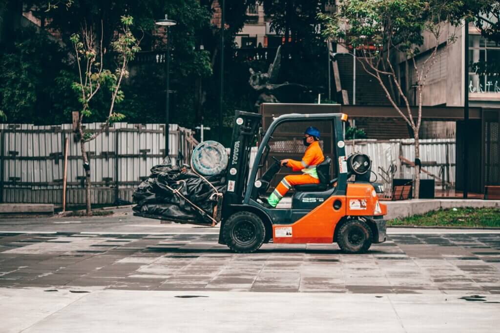 Een persoon in een oranje uniform bestuurt vakkundig een oranje vorkheftruck, die zwarte zakken en een blauw vat vervoert. Terwijl ze over het geplaveide gebied rijden met bomen en gebouwen op de achtergrond, zou men de voordelen van een groepstrainingssessie voor zon bekwame operatie kunnen overwegen.