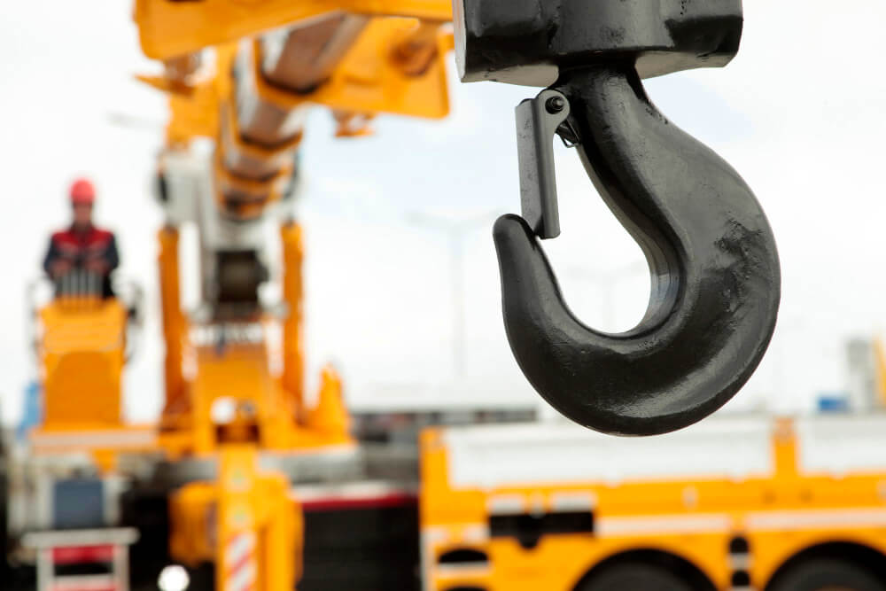 Close-up van een grote zwarte kraanhaak in focus, met een wazige achtergrond waarop een bouwvakker in een hoogwerker en de gele kraanbody te zien zijn. Geïnteresseerd in het leren van veilige hijstechnieken op locatie? Cursus veilig hijsen bij jullie op locatie biedt meerdere mogelijkheden.