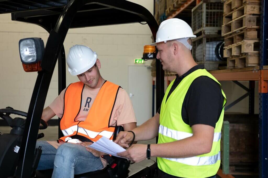 Twee arbeiders in een magazijn, tussen de stellingen met dozen, bespreken documenten. De een zit op een heftruck in een oranje vest en helm en bekijkt de papieren, terwijl de ander, staand in een geel vest, naar hen wijst. De herfst is begonnen. Profiteer van onze herfstactie op valbeveiliging!.