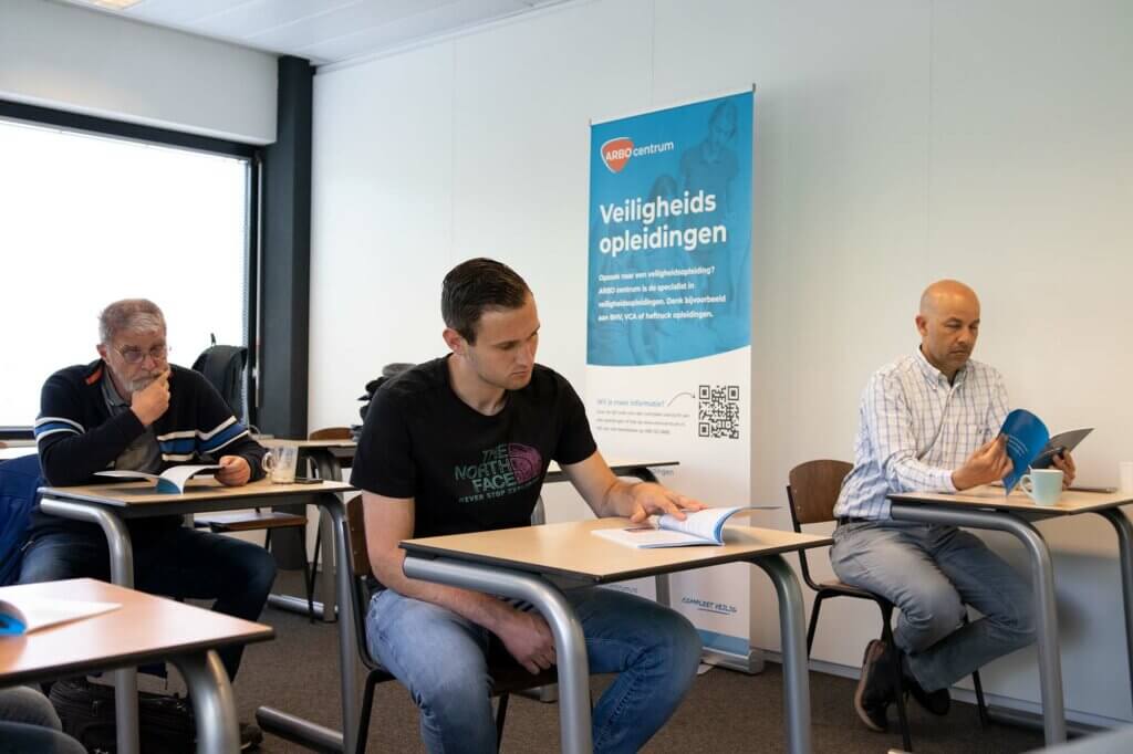 Drie mannen zitten aan een bureau in een klaslokaal en lezen uit boekjes. Op een spandoek achter hen staat Veiligheidsopleidingen. De kamer heeft een licht raam en een modern interieur. De eerste VCA cursus Arabisch is een feit!.