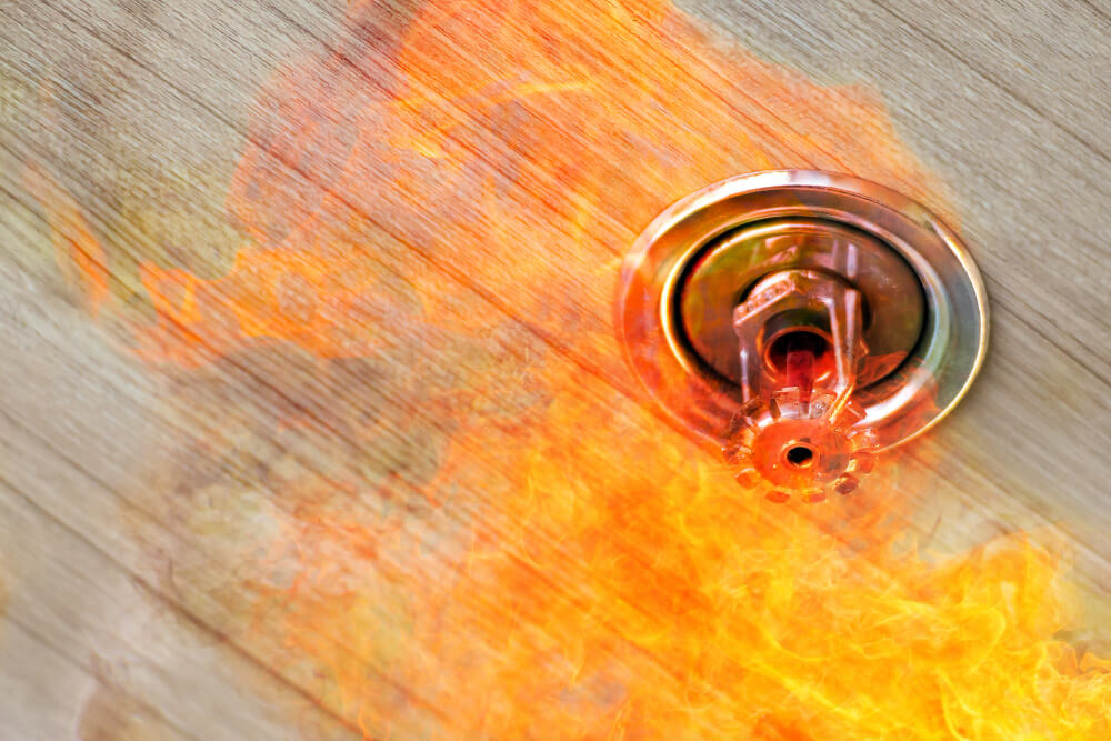 Een sprinklerinstallatie op een houten plafond wordt geactiveerd en laat water vrijkomen te midden van felle, oranje vlammen. Terwijl het vuur zich verspreidt en De Nationale Brandpreventieweken op 1 oktober beginnen, is het sprinklersysteem cruciaal bij het beheersen van de brand.
