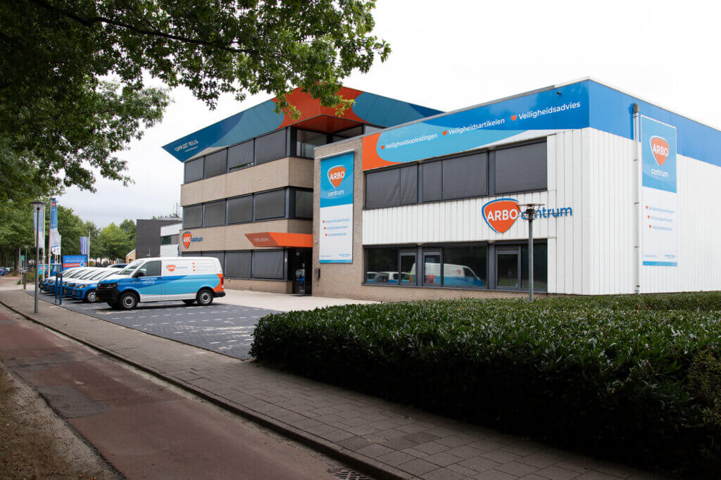 Het moderne kantoorgebouw heeft een strak wit en blauw exterieur met grote ramen met daarop borden voor ARBO centrum. Merkvoertuigen, die onder specifieke actievoorwaarden vallen, staan geparkeerd op het terrein, omgeven door weelderige bomen en groen. Een trottoir nodigt gasten uit om het terrein te verkennen.