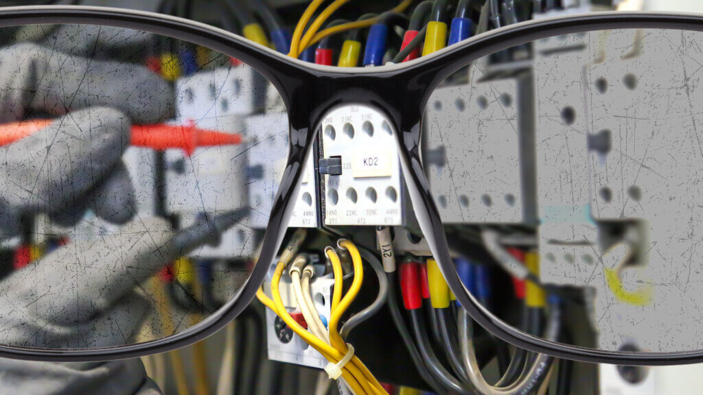 Close-up van een bril die focust op een elektrisch paneel met draden en schakelaars. Ter ere van De internationale dag van het zicht, is een persoon met handschoenen bezig met het aanpassen van een onderdeel met een schroevendraaier. Het montuur van de bril is prominent op de voorgrond, waardoor het zicht gedeeltelijk wordt belemmerd.