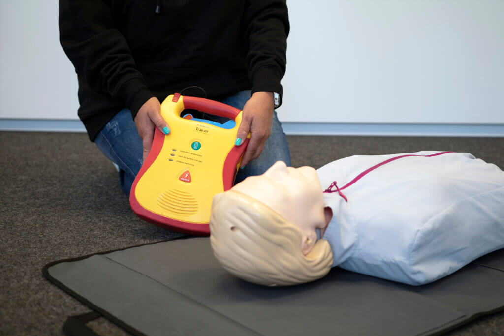 Op World Restart a Heart Day demonstreert een persoon het gebruik van een automatische externe defibrillator (AED) op een reanimatie-oefenpop die op een mat ligt. De AED, felgeel met rode randen, helpt bij het aanleren van levensreddende vaardigheden terwijl de pop gekleed is in een wit jasje.
