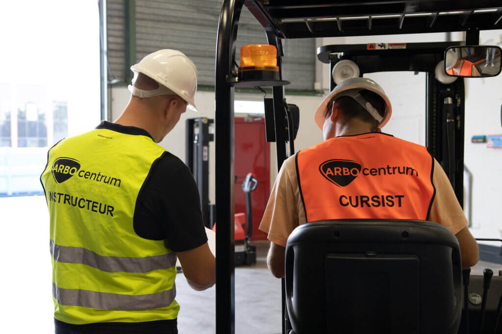 Twee mensen met helmen en veiligheidsvesten worden van achteren gezien naast een vorkheftruck tijdens De dag van de leraar. Op het vest links staat Instructor en op het vest rechts staat Cursist. Ze lijken zich in een magazijn of industriële omgeving te bevinden.
