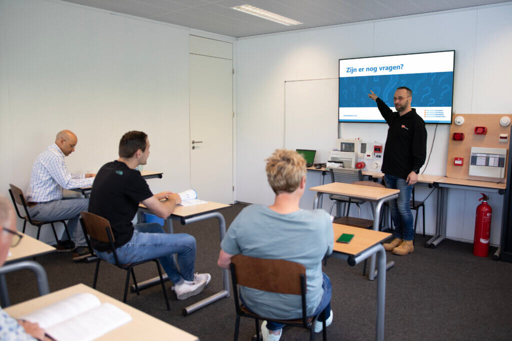 Een leraar staat vooraan in een klaslokaal en wijst naar een groot scherm met tekst over hoe een veilige werkplek de productiviteit van werknemers verhoogt. Vier studenten zitten aan bureaus, aandachtig betrokken. Het klaslokaal toont brandveiligheidsapparatuur en een brandblusser.