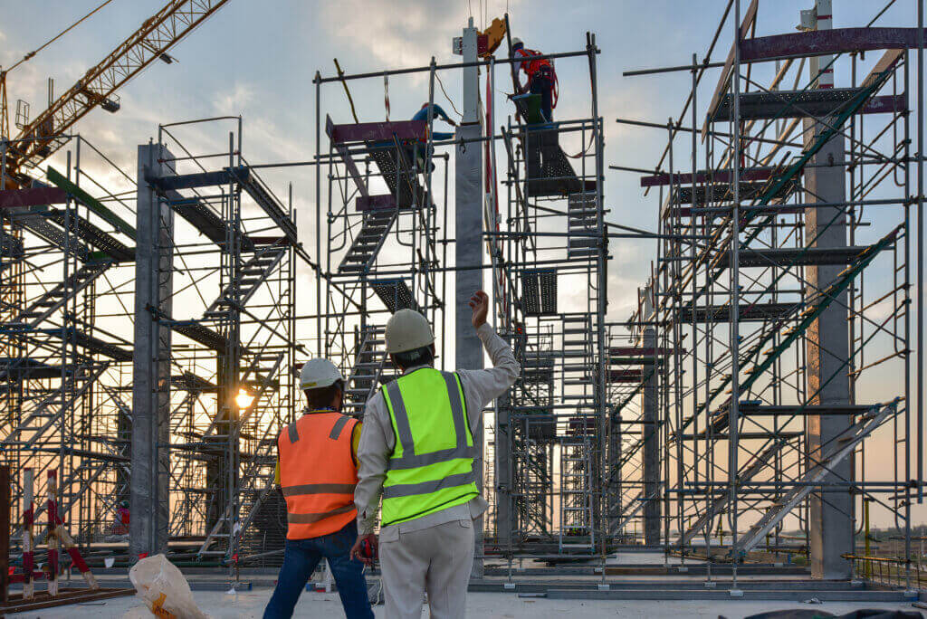 Construction workers on a building site with scaffolding. Two workers wearing safety vests and helmets discuss plans while others work on elevated scaffolding. A crane is visible in the background against a cloudy sky.