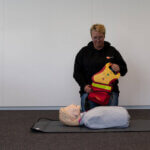 A person is kneeling in a room, participating in an AED cursus as they prepare to use a defibrillator on a CPR training mannequin lying on a mat. The mannequin, with a blond head and blue top, awaits the next step in this vital life-saving exercise.