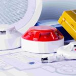 Various fire alarm and safety devices, including smoke detectors and a yellow manual call point, are placed on top of blueprints. A cable and a circular speaker suggest planning or installation work, possibly as part of a brand en ontruiming cursus.