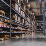 A large warehouse aisle with tall metal shelves filled with various-sized boxes and wooden pallets showcases a pristine floor. The organized environment reflects the precision learned in an orderpicktruck cursus, ensuring efficiency in every corner of the storage space.