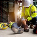 A construction worker in a high-visibility jacket and helmet, who recently completed a preventiemedewerker cursus, attends to a colleague lying on the ground, grabbing their knee. A yellow helmet is beside the injured person, and a red first aid bag is nearby on the busy construction site with scaffolding.
