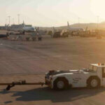 A sunny airport tarmac scene unfolds with several parked airplanes in the background. In the foreground, a white pushback tug vehicle, reminiscent of a Terminaltrekker minus its operator, stands poised. The sky is clear, graced with a warm glow from the sun.