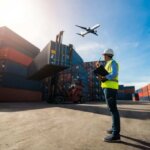 A worker in a hard hat and reflective vest stands with a clipboard, observing stacked shipping containers and a forklift. With an airplane flying overhead in the clear sky, it suggests a busy logistics scene, enhanced by the precision learned from a Zijlader cursus.