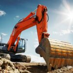 Een grote oranje graafmachine, die lijkt op een veelzijdige grondverzet minigraver, staat geparkeerd op ruw, rotsachtig terrein onder een helderblauwe lucht. De zon schijnt fel op de achtergrond en werpt schaduwen op de grond terwijl de arm en bak prominent worden weergegeven.